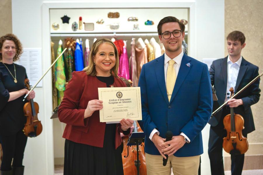 Two adults stand indoors holding a framed “Certificate of Congressional Recognition and Achievement” while several musicians with string instruments stand behind them; one person holds a microphone. The background shows a display of colorful garments or costumes on racks within a museum or gallery setting.