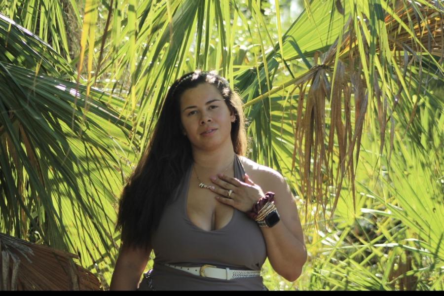 Waist up shot of Siera standing against background of palm trees. She has long brown hair and wears an army green tank top