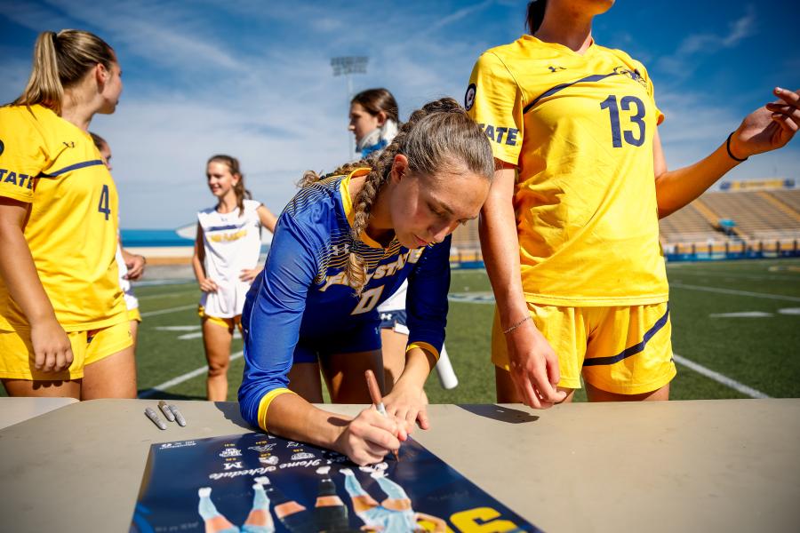 Emma Klein signing a poster on the field while surrounded by fellow team mates. 