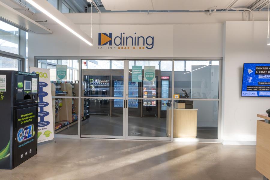 Front view of the DI Dining Grab-n-Go area, featuring closed sliding-glass doors. Inside the area are racks of snacks, display coolers, and a check-out counter.