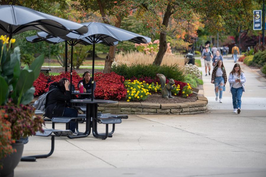Students walking and sitting near outdoor tables, flower beds and a squirrel sculpture on campus.