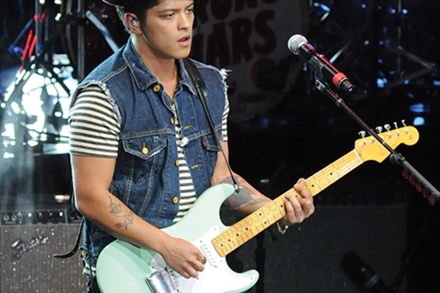 Bruno Mars playing electric guitar at the MAC Center, wearing a striped shirt, denim vest, and fedora.