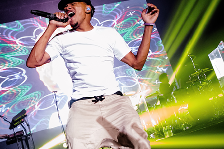 Chance the Rapper performing at the MAC Center, wearing a white shirt and black cap while holding a microphone on stage.