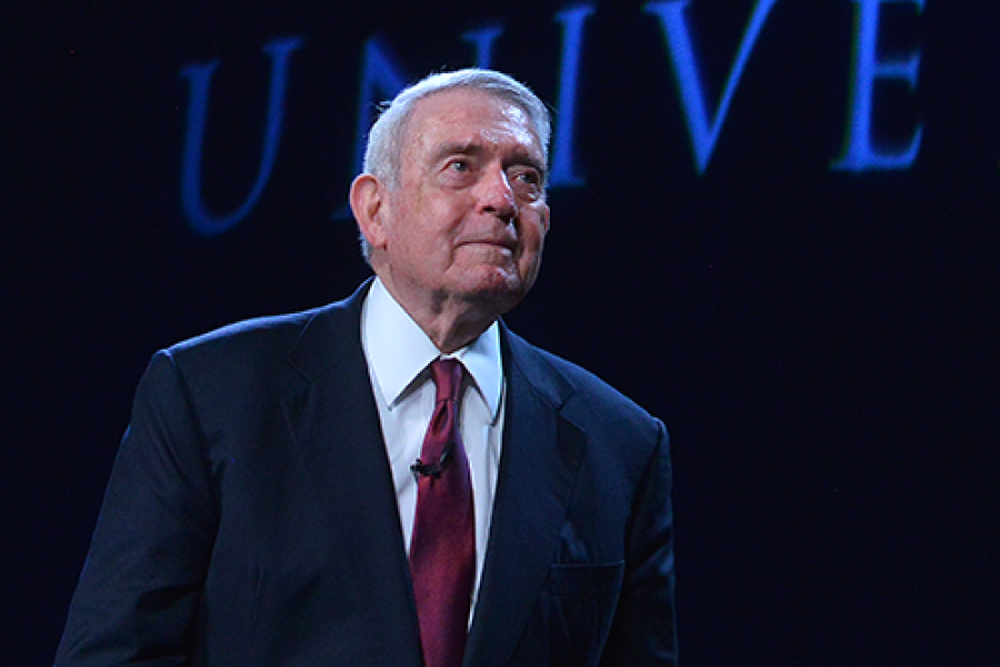 Dan Rather speaking at the MAC Center, wearing a dark suit and red tie with a university backdrop behind him.