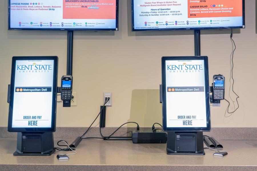 Front view of the order station at Metropolitan Deli, featuring menu screens on the wall above and two ordering kiosks