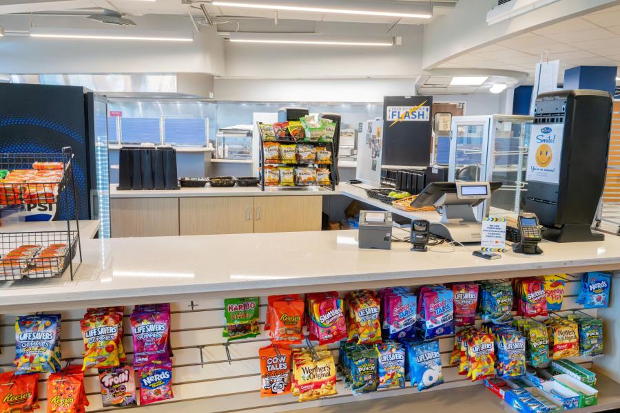 Checkout counter at Rosie's Market, including a register, payment device, snack options displayed, and a napkin dispenser