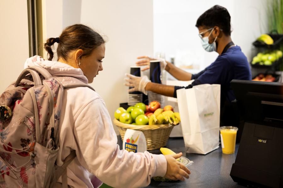 A female student picking up her order at Recharge's pick-up station. A basket of fresh fruit is on the counter and the student employee is grabbing a fountain drink cup.