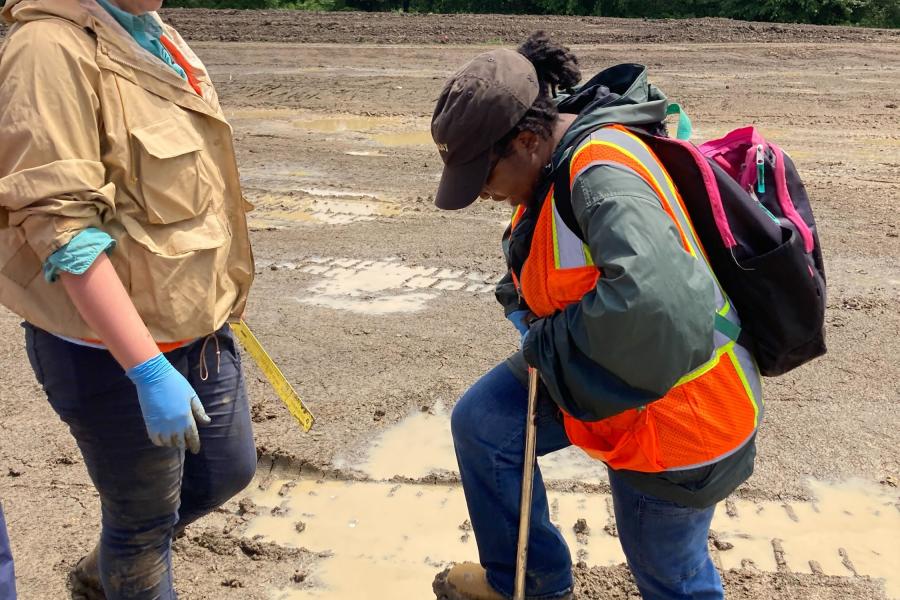 A woman is digging in the dirt with a shovel, wearing a hi-vis vest. Another woman stands in front of her, supervising, and wearing a hard hat. They are both in a barren landscape, there are trees far off in the background.