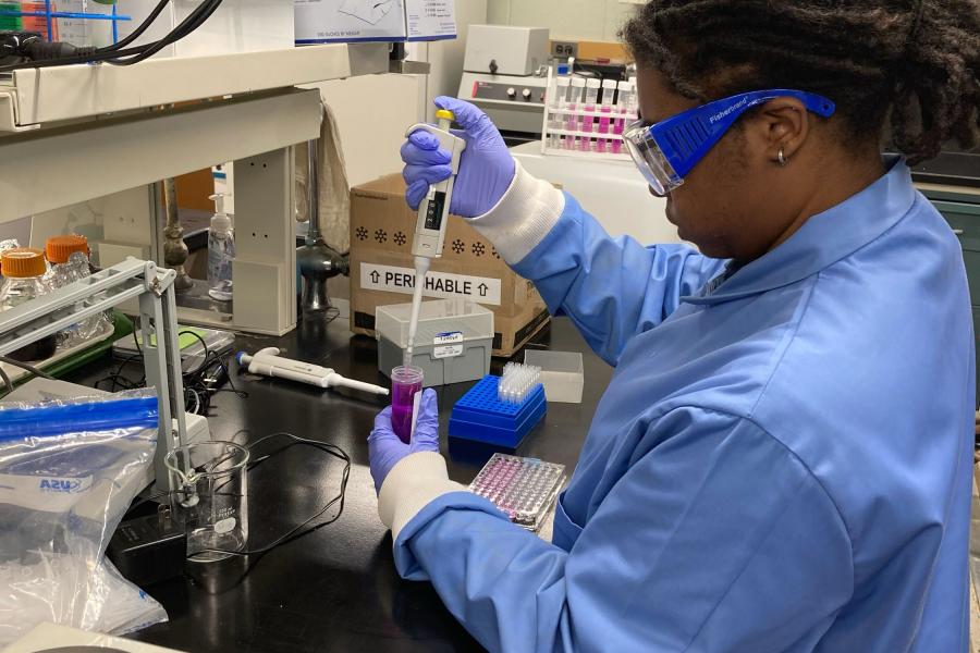 A woman is in a lab, wearing a lab coat and goggles. She is transferring a liquid from a pipette to a beaker.