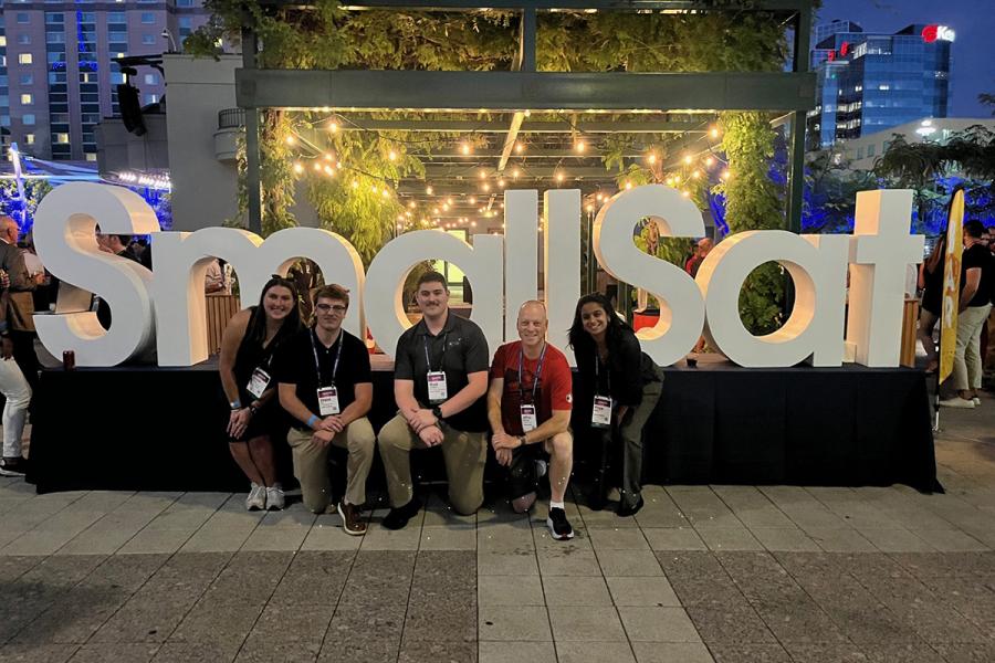 a small group of students standing in front of a large sign that reads "SmallSat"