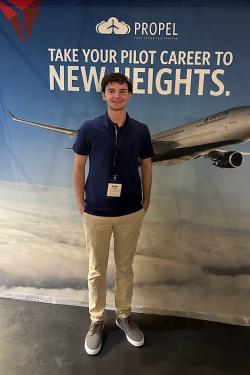 A young man in a blue t-shirt stands in front of a large Delta Propel recruitment poster