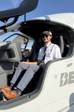 A young man wearing a dress shirt and tie sits in the cockpit of a modern electric aircraft
