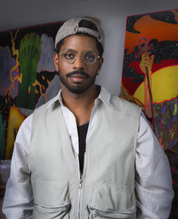 A man with a backwards beige cap and round glasses stands with his arms by his sides for a portrait, pictured from the waist up. He wears a beige vest and white long sleeve shirt underneath