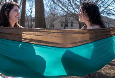 Two students sit in a hammock outside on a sunny day