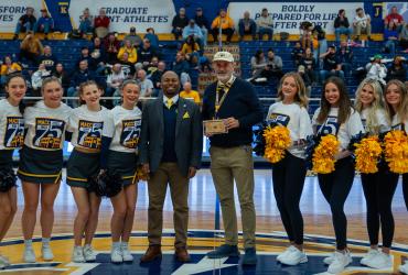 Group of cheerleaders and three adults smiling and posing at a sports event, holding an award, with spectators in the background. The group includes individuals in cheer uniforms and others in formal attire.
