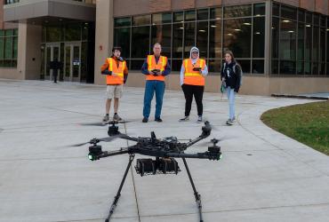 Four people standing at a distance wearing high-visibility vests with a medium-sized quadcopter in the foreground.
