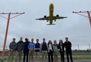 Philip standing with fellow aeronautics students at the airport with a plane flying above