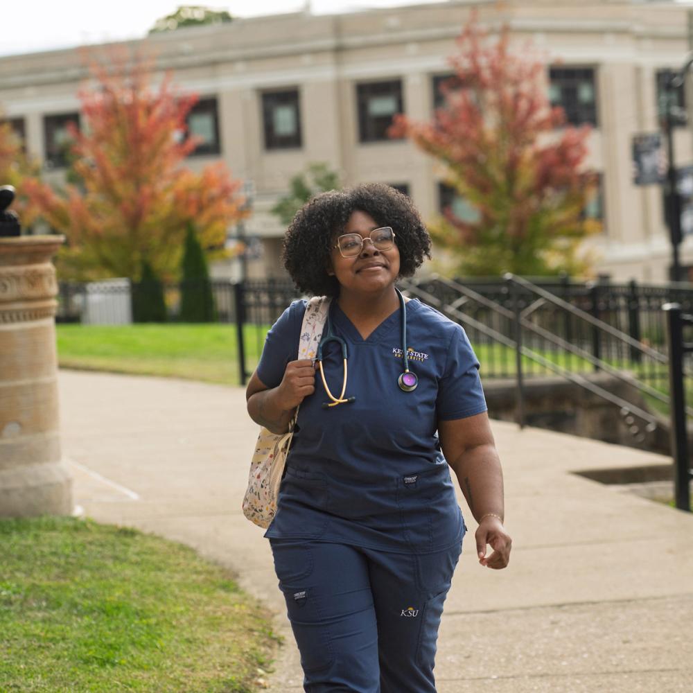A nursing student wearing scrubs and a stethoscope walks across the campus.