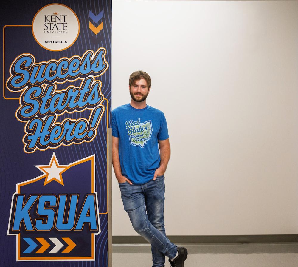 A man wearing a blue 51������ Regional Campuses T-shirt stands beside a 51������ Ashtabula “Success Starts Here” banner.