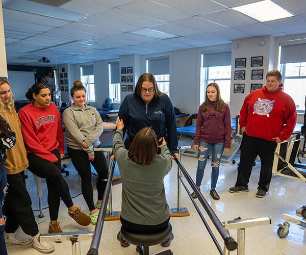 Physical therapist assistant students observe a seated mobility demonstration using parallel bars in a classroom.