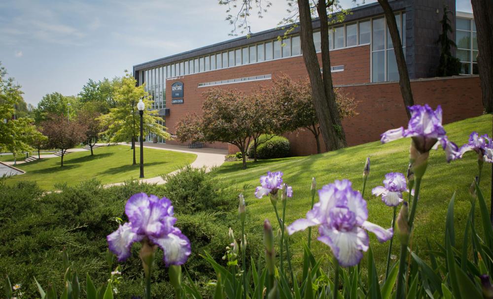 The Campus Center is surrounded by landscaped lawns, trees, and blooming purple flowers on a sunny day.