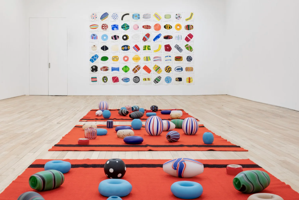 Photograph of a contemporary art installation featuring colorful, patterned spherical and cylindrical glass objects arranged on red fabric strips laid out on a wooden floor. A large grid of similarly styled, vibrant, abstract shapes is displayed on a white wall in the background, creating a cohesive visual theme of geometric forms and bold colors.