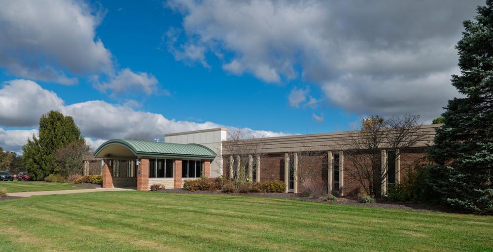 A brick campus building with a curved green entrance canopy under a partly cloudy sky.