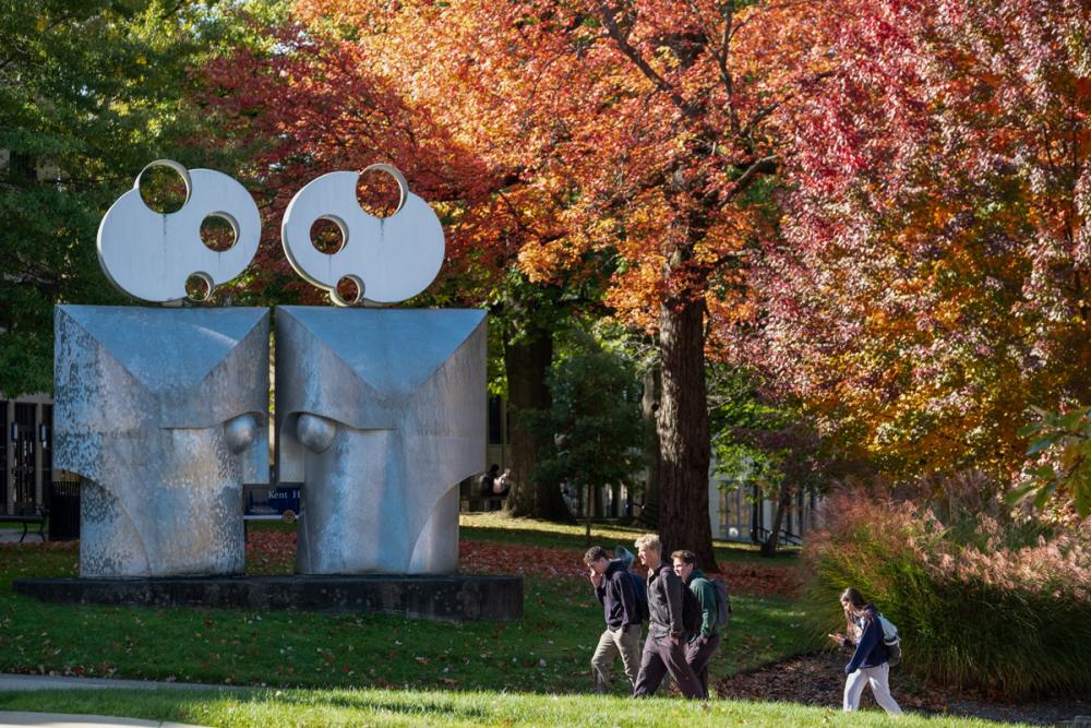 Students walking past an outdoor sculpture surrounded by vibrant fall foliage.