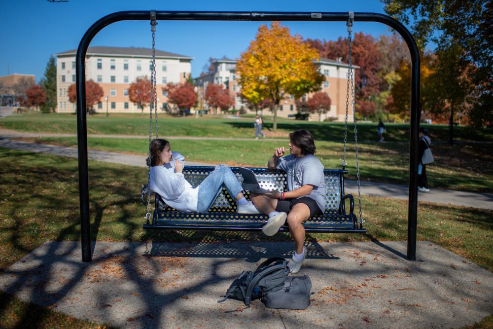 Two students talking while seated on a swing bench on campus.