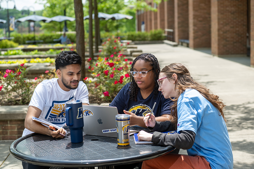 Three students sit at a table outside on campus.