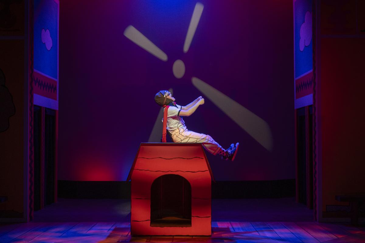 A student performer dressed as Snoopy sits atop a red doghouse, leaning back with arms extended, in a theatrical stage setting with vibrant lighting and stylized backdrops.