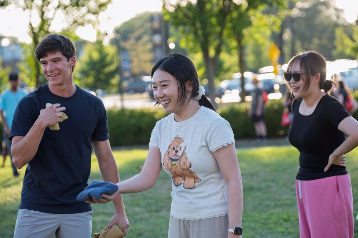 Three students play corn hole. The student in the center holds a bean page and takes a underhanded throwing motion with two others flank her and smile.