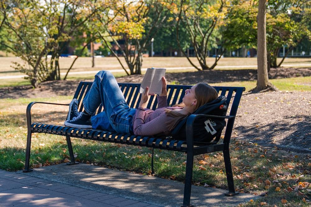 Student reads a book and lays on a bench outside