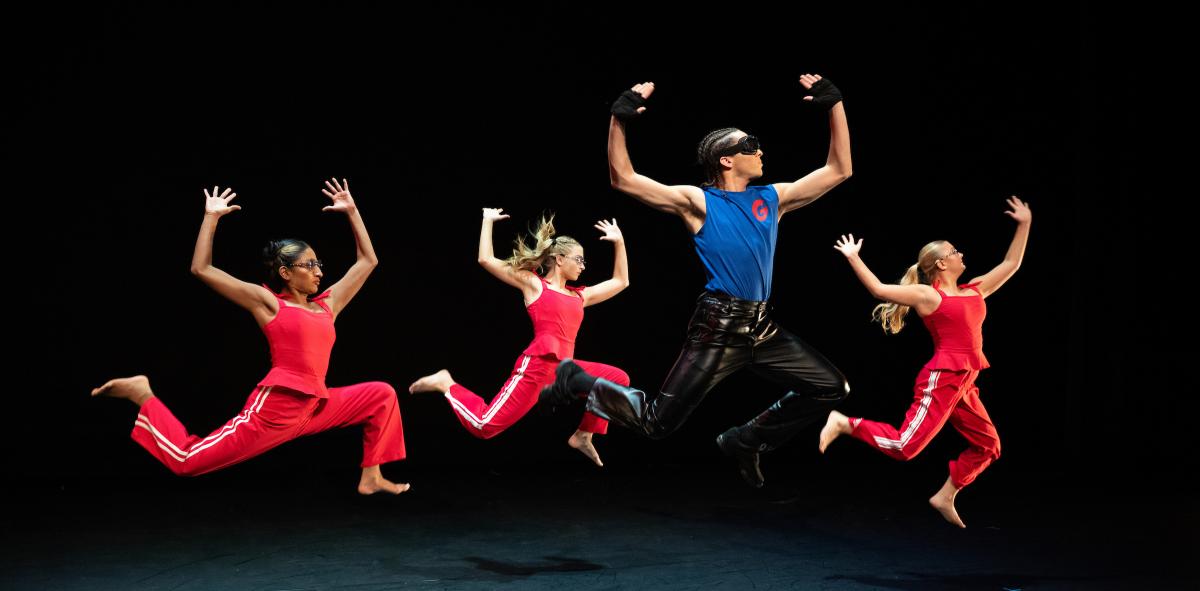 Performers on stage, featuring three in red attire and one in a blue and black outfit, all engaged in a dynamic dance routine.