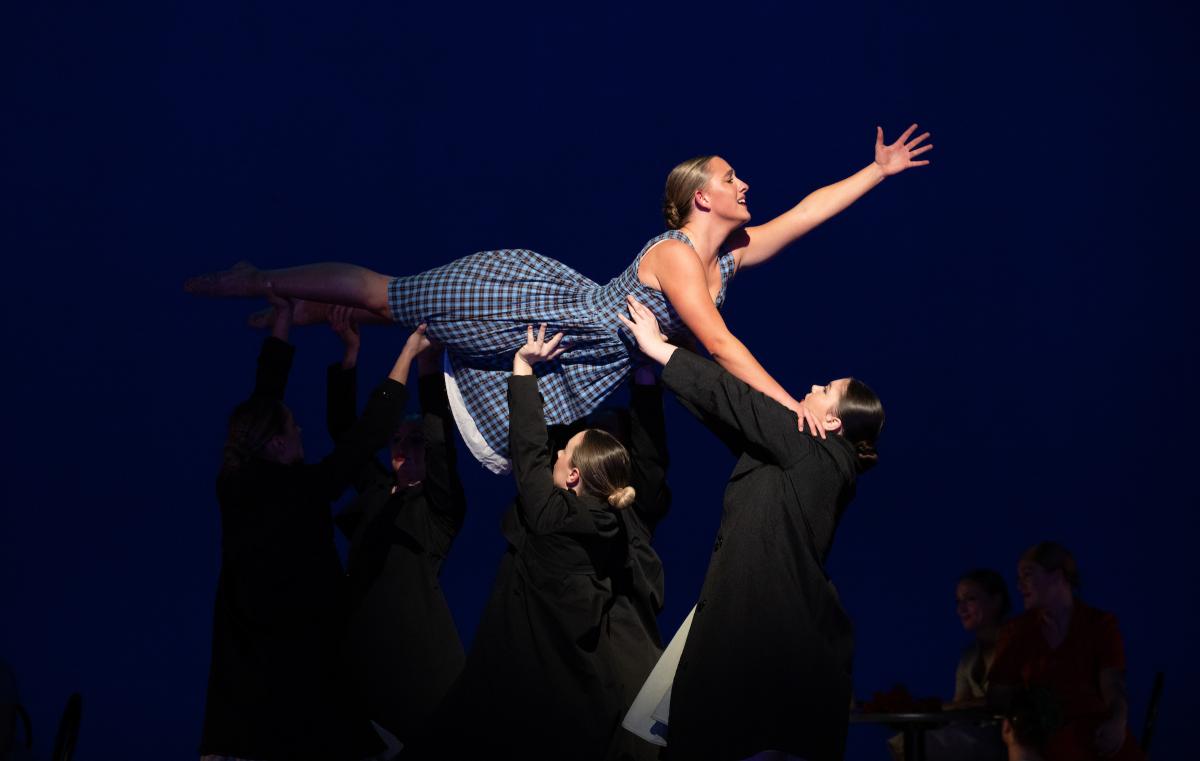 A performer is lifted horizontally by a group of dancers in dark suits against a blue backdrop; the central figure is smiling, arms outstretched, wearing a plaid dress.