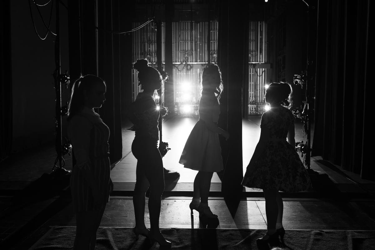Four silhouetted individuals standing on a stage, backlit by bright stage lights, creating a dramatic effect.