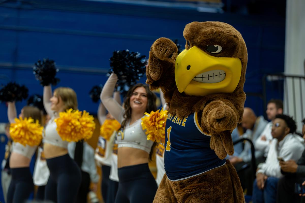 ɫҹ University mascot, a person dressed in an oversized eagle costume with a happy expression, is surrounded by cheerleaders holding pom-poms at a sports event.