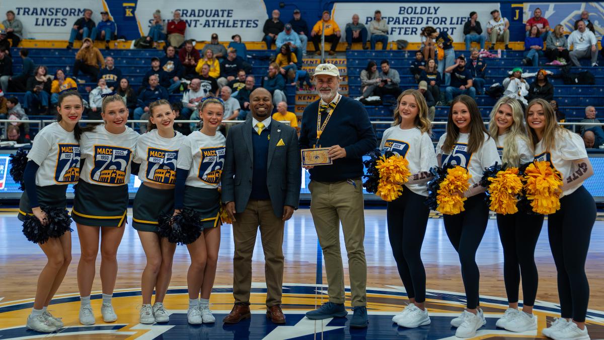 Group of cheerleaders and three adults smiling and posing at a sports event, holding an award, with spectators in the background. The group includes individuals in cheer uniforms and others in formal attire.