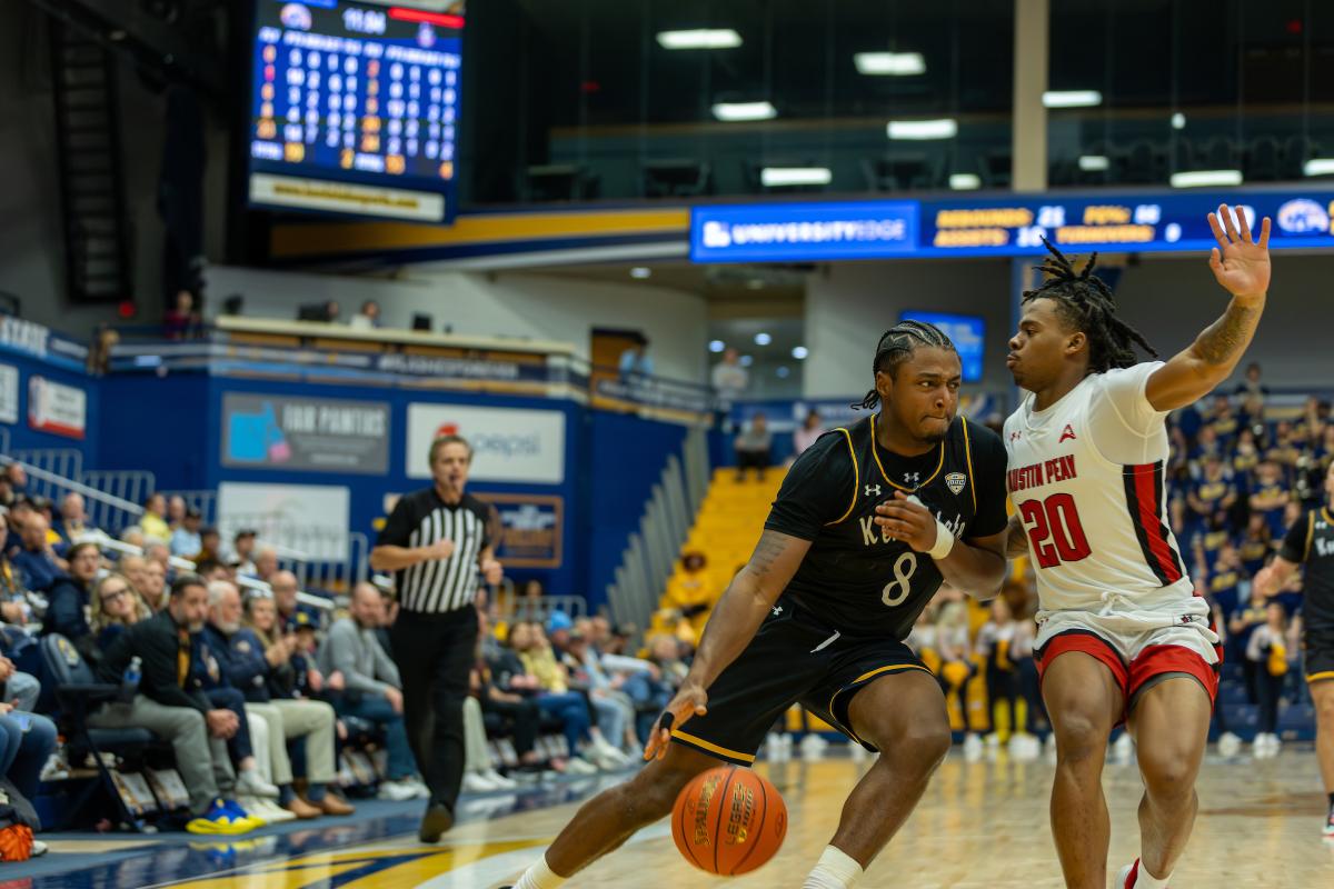 Two basketball players from ɫҹ and Austin Peay are in action during a game at a crowded arena, with one dribbling the ball and the other defending. The scoreboard in the background shows scores and game information.