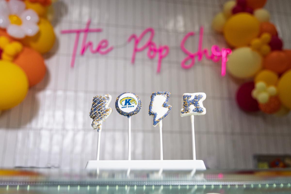 Four white chocolate-dipped cake pops shaped like a lightning bolt, a circular �Ը������� logo, a lightning bolt outline and the letter "K," decorated in blue and gold sprinkles, displayed on a stand in front of Daisy Pops' pink neon "The Pop Shop" sign.