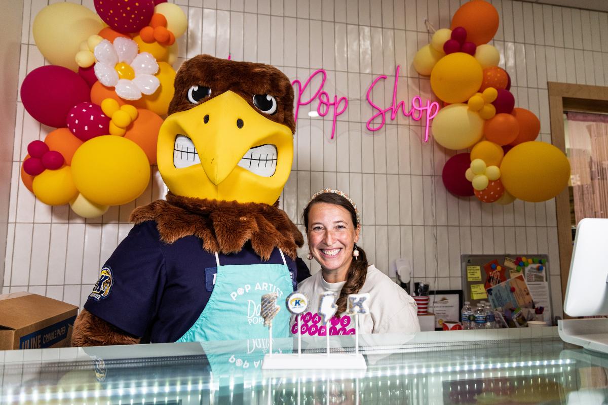 Flash the �Ը������� mascot poses with a smiling Daisy Pops owner behind the shop counter, with four �Ը�������-themed cake pops on display and a balloon garland and neon "Pop Shop" sign behind them.