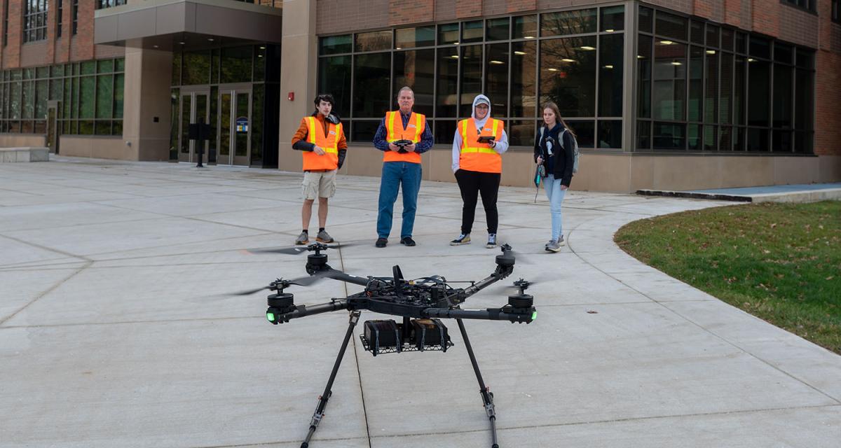 Four people standing at a distance wearing high-visibility vests with a medium-sized quadcopter in the foreground.