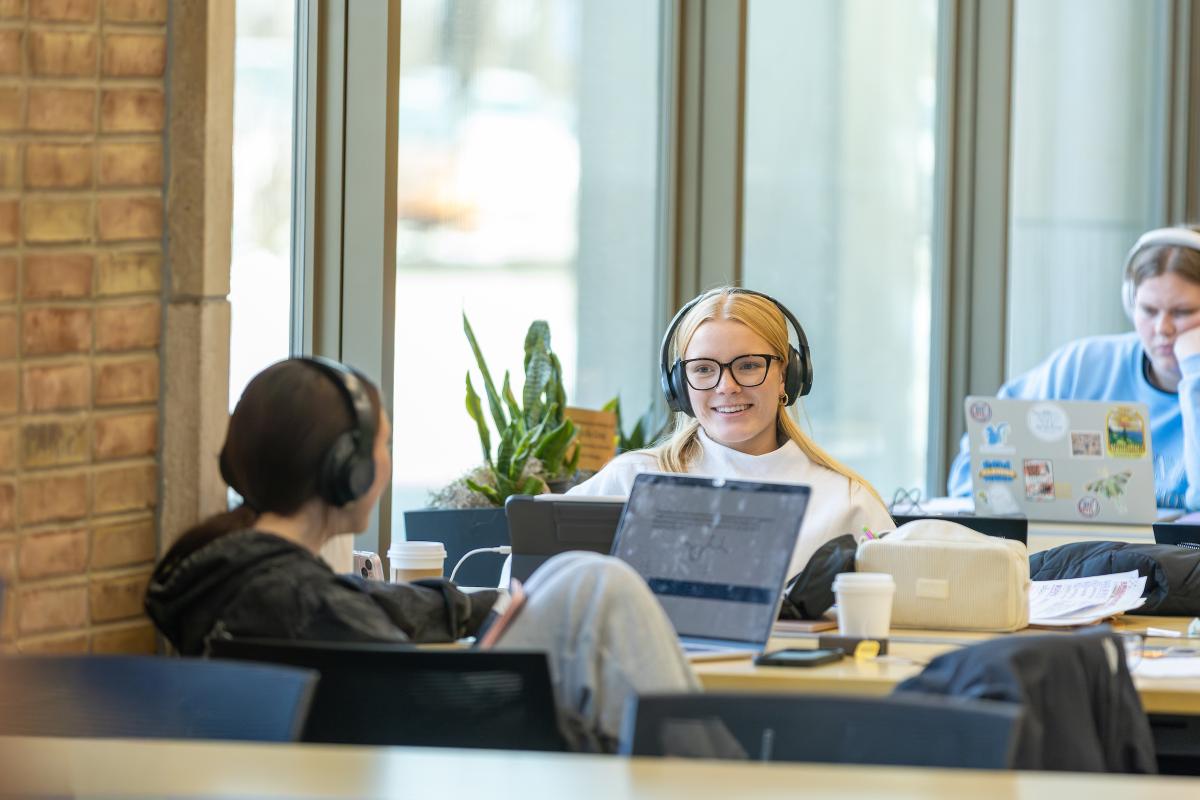 Person in headphones smiling at laptop in a public space, surrounded by other people working.