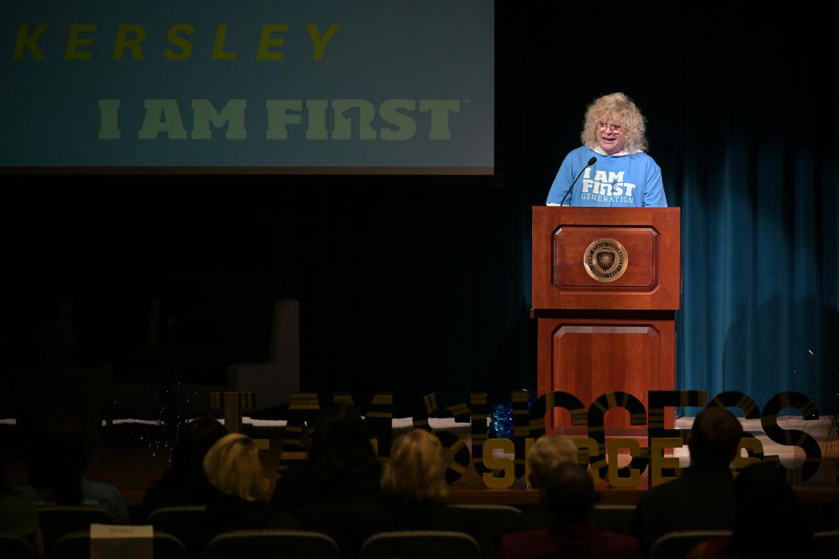 鶹ѡProvost and keynote speaker stands at a podium with a banner reading "I AM FIRST" in a lecture hall, addressing an audience. The background features a large screen with text.