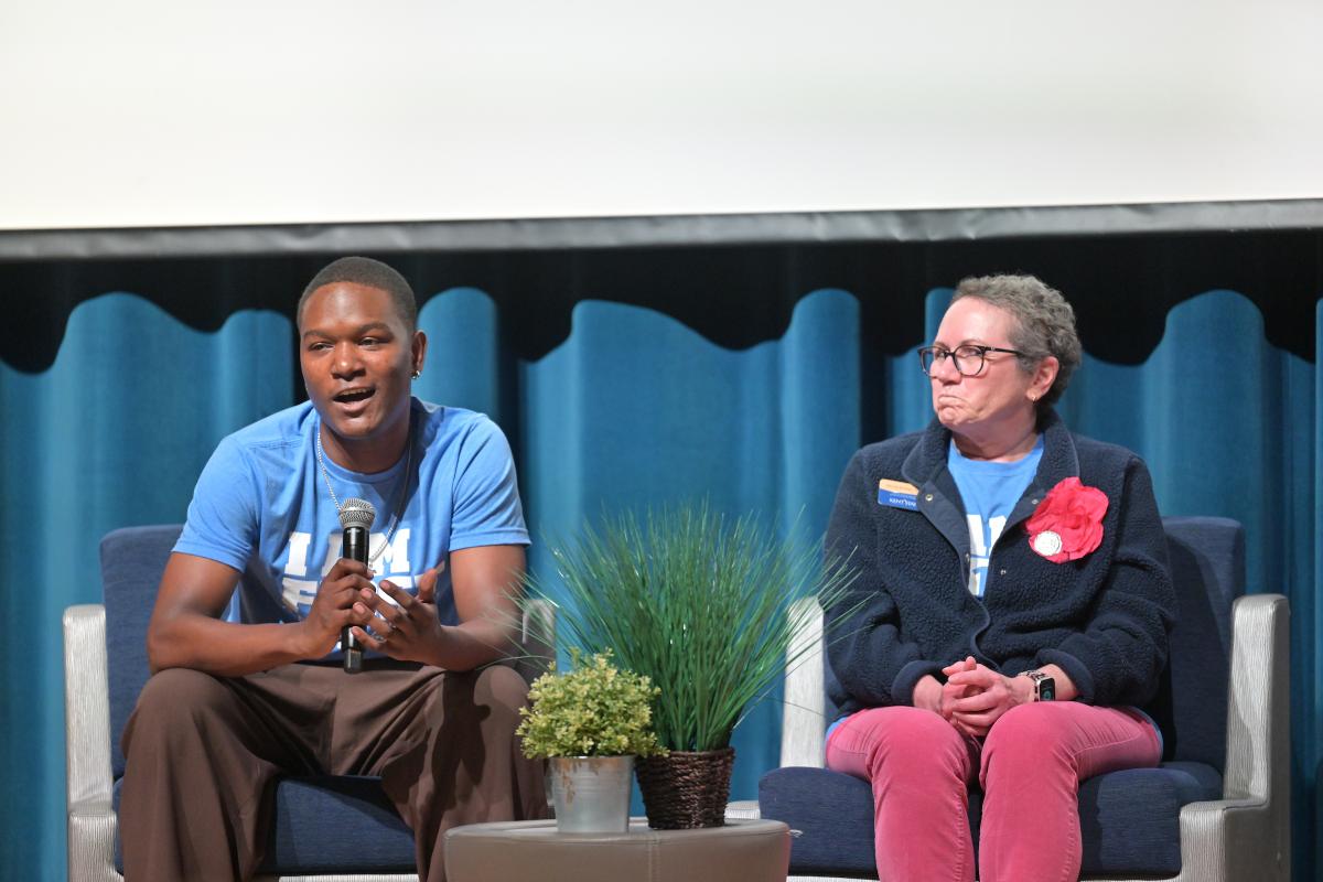 Two people participating in a panel discussion, one speaking with a microphone and the other listening attentively. There's a potted plant on the stage between them.