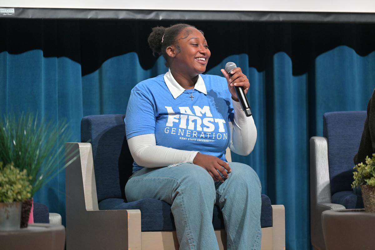Person sitting on a stage, holding a microphone and wearing a blue 'I AM FIRST GEN' t-shirt, smiling during a discussion at an event.