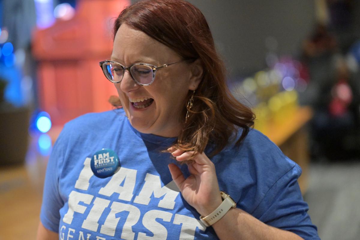 A person wearing glasses and a blue "I AM FIRST" shirt laughs joyfully at an event.