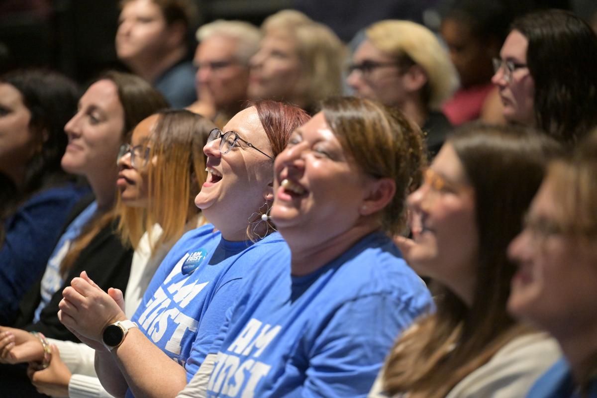 Audience members in blue shirts, some with I AM FIRST logos, laughing and enjoying a presentation.