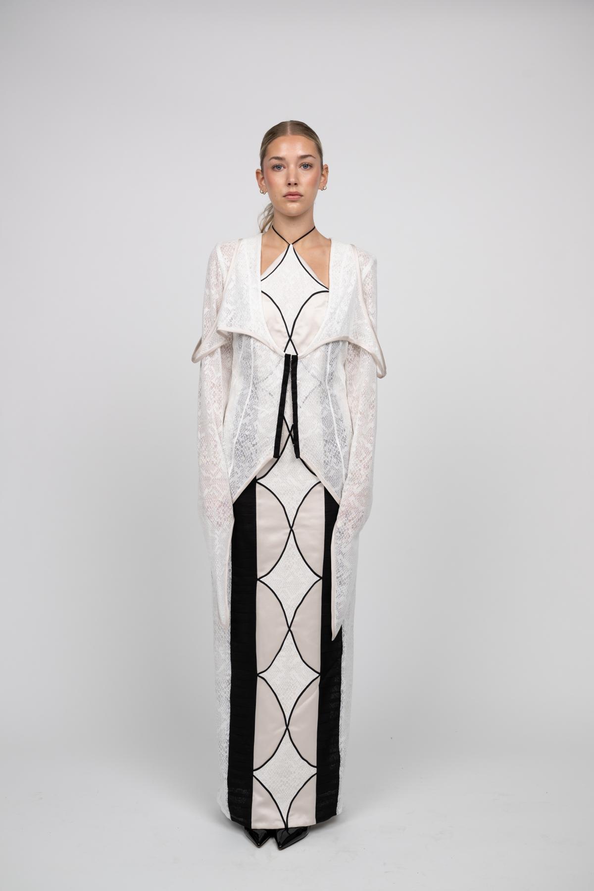 a model wears a symmetrical black and white gown resembling a stained glass window. a knitwear shawl is worn over top 