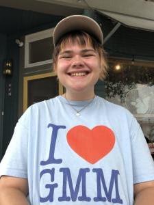 Finn Machuga smiles while standing outdoors in a light blue "I Love GMM" T-shirt and cap in front of a building entrance.